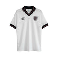 Camiseta de fútbol retro blanca de manga corta de Umbro para Inglaterra 1984, con cuello en V negro y ribete rojo, y escudo del equipo bordado.